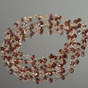 Può includere: Una delicata collana con piccole perline sfaccettate marrone-rossastre infilate su filo dorato. Le perline sono uniformemente distanziate, creando un effetto a strati. Il gioiello è esposto su una superficie riflettente.