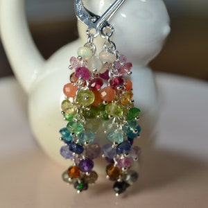 May include: A pair of dangle earrings with a rainbow of gemstones. The earrings feature a silver leverback clasp and multiple strands of colorful beads, including pink, red, orange, yellow, green, blue, and purple. The gemstones are small and round.