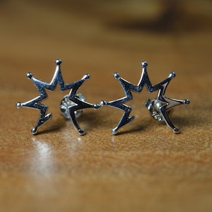 以下が含まれることがあります： 銀色のスタッドピアス。各イヤリングは、尖った先端と中央のオープンスペースを備えた、様式化されたスターバーストデザインが特徴です。イヤリングは木製の表面に表示されています。