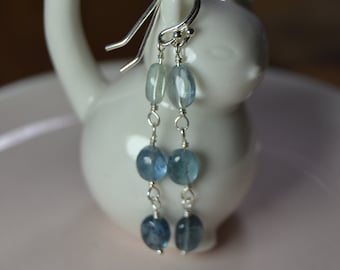 May include: Dangling earrings with blue and light blue beads. The earrings are suspended from silver-coloured hooks and are displayed on a white cat-shaped ceramic holder. The earrings have a simple, elegant design.