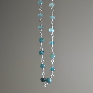 May include: A delicate necklace featuring small, round, blue-green gemstones strung on a silver chain. The gemstones vary slightly in shade, creating a subtle gradient effect. The necklace is displayed against a neutral gray background.