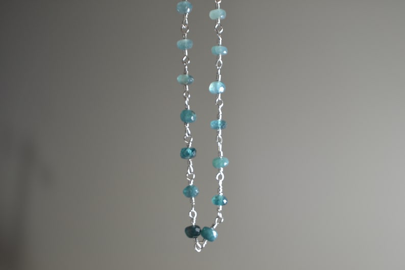 May include: A delicate necklace featuring small, round, blue-green gemstones strung on a silver chain. The gemstones vary slightly in shade, creating a subtle gradient effect. The necklace is displayed against a neutral gray background.