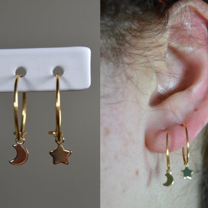 Puede incluir: Pendientes de aro dorados con colgantes celestiales. Un pendiente presenta un dije de media luna, el otro una estrella. Los pendientes se muestran en un soporte blanco y se usan en una oreja.