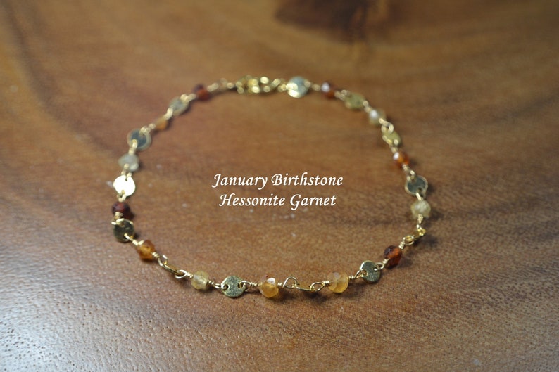 May include: A delicate gold-toned bracelet featuring small, round gold accents and various-sized, colorful gemstones. The text "January Birthstone Hessonite Garnet" is displayed on the image. This piece is likely a jewelry item.