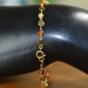 May include: Gold-toned bracelet featuring small, round, gold-colored discs and orange-brown beads. The bracelet is connected by a delicate gold chain and has a clasp closure. The jewelry is displayed on a black surface.
