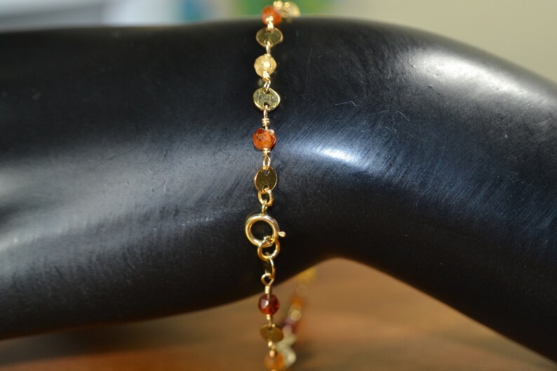 May include: Gold-toned bracelet featuring small, round, gold-colored discs and orange-brown beads. The bracelet is connected by a delicate gold chain and has a clasp closure. The jewelry is displayed on a black surface.