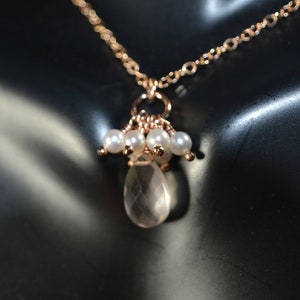 Puede incluir: Un delicado collar de oro rosa con una fina cadena, adornado con un grupo de peque&ntilde;as perlas blancas y un colgante de cuarzo transparente en forma de l&aacute;grima. Se muestra sobre un fondo oscuro.
