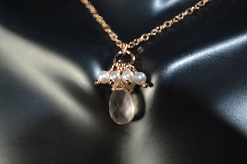 Puede incluir: Un delicado collar de oro rosa con una fina cadena, adornado con un grupo de peque&ntilde;as perlas blancas y un colgante de cuarzo transparente en forma de l&aacute;grima. Se muestra sobre un fondo oscuro.
