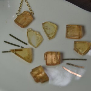 May include: A collection of amber-colored crystal pendants and loose crystals on a white surface. Two necklaces with gold and silver chains are visible. The crystals vary in shape, with some appearing rectangular and others more irregular.