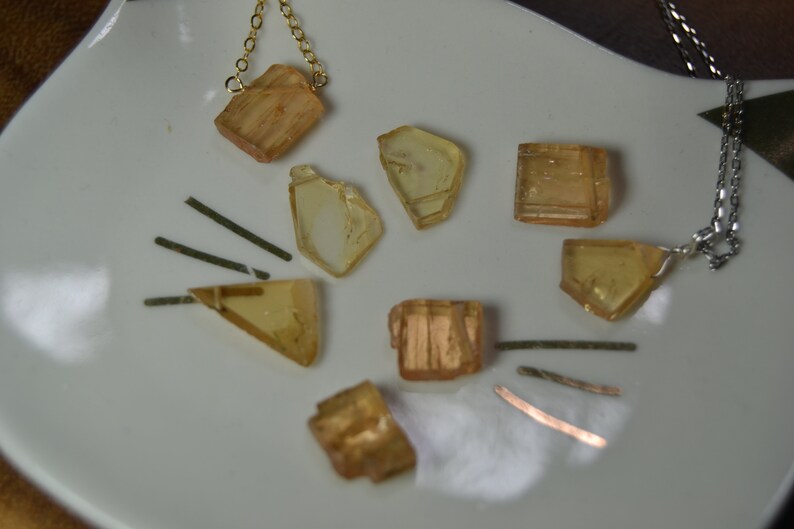 May include: A collection of amber-colored crystal pendants and loose crystals on a white surface. Two necklaces with gold and silver chains are visible. The crystals vary in shape, with some appearing rectangular and others more irregular.