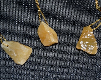 May include: Three necklaces with raw, amber-colored stone pendants. Each stone is irregularly shaped and suspended from a delicate gold-colored chain. The background is a dark, textured surface.