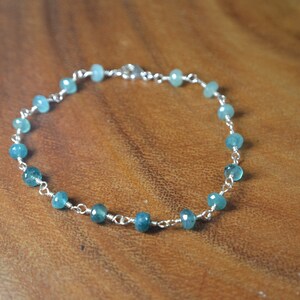 May include: A delicate bracelet featuring small, faceted, teal-colored beads strung on a silver-colored chain. The beads are evenly spaced along the chain, creating a simple yet elegant design. The bracelet is displayed on a wooden surface.