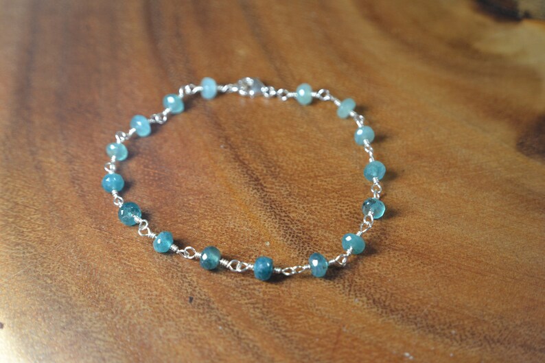 May include: A delicate bracelet featuring small, faceted, teal-colored beads strung on a silver-colored chain. The beads are evenly spaced along the chain, creating a simple yet elegant design. The bracelet is displayed on a wooden surface.