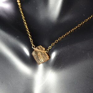 May include: A delicate gold-colored necklace featuring a small, rough-cut, amber-colored gemstone pendant. The chain is thin and the pendant is rectangular. The necklace is displayed on a black surface.