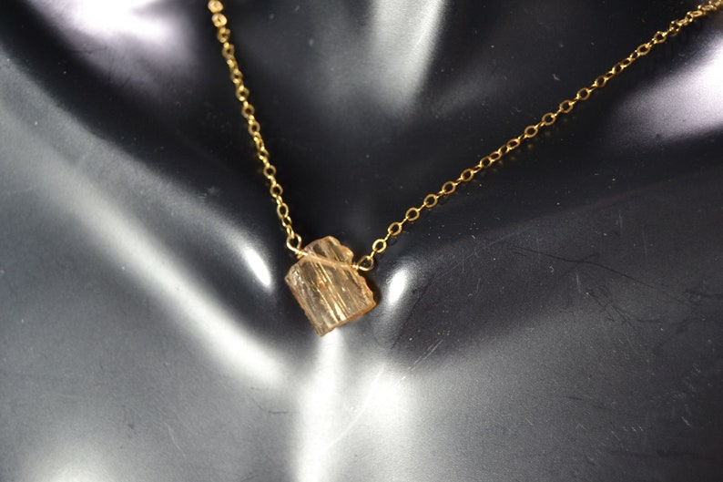 May include: A delicate gold-colored necklace featuring a small, rough-cut, amber-colored gemstone pendant. The chain is thin and the pendant is rectangular. The necklace is displayed on a black surface.