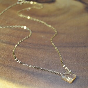 May include: A delicate gold-colored necklace with a small, square-shaped pendant. The pendant appears to be a light amber color. The necklace chain is thin and has a simple, elegant design. The necklace is displayed on a wooden surface.
