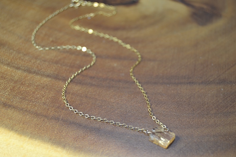 May include: A delicate gold-colored necklace with a small, square-shaped pendant. The pendant appears to be a light amber color. The necklace chain is thin and has a simple, elegant design. The necklace is displayed on a wooden surface.