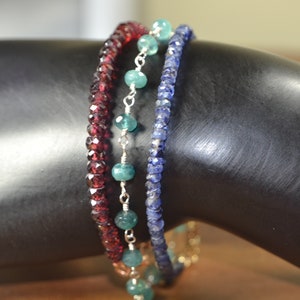 May include: Three beaded bracelets in shades of red, teal, and blue. The red bracelet features small, faceted beads. The teal bracelet has larger, round beads strung on a silver chain. The blue bracelet is made of small, faceted beads.