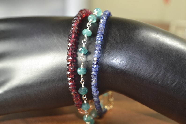 May include: Three beaded bracelets in shades of red, teal, and blue. The red bracelet features small, faceted beads. The teal bracelet has larger, round beads strung on a silver chain. The blue bracelet is made of small, faceted beads.