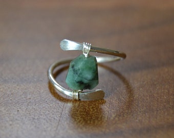 May include: A silver-toned ring with a dark green gemstone. The ring features a spiral design with a rectangular gemstone pendant. The gemstone is a deep green color with visible inclusions. The ring is set against a brown wooden surface.