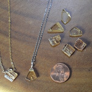 May include: Two necklaces with clear, amber-colored crystal pendants, alongside loose crystals and a penny for scale. One necklace has a gold chain, the other a silver chain. The crystals have a rough, natural appearance, and are set against a brown background.