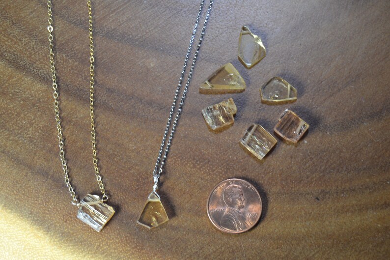 May include: Two necklaces with clear, amber-colored crystal pendants, alongside loose crystals and a penny for scale. One necklace has a gold chain, the other a silver chain. The crystals have a rough, natural appearance, and are set against a brown background.
