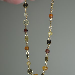 May include: Delicate gold necklace featuring a series of small, colorful gemstones and gold-colored disc accents. The gemstones vary in shades of amber, brown, and yellow, strung together with fine gold wire.