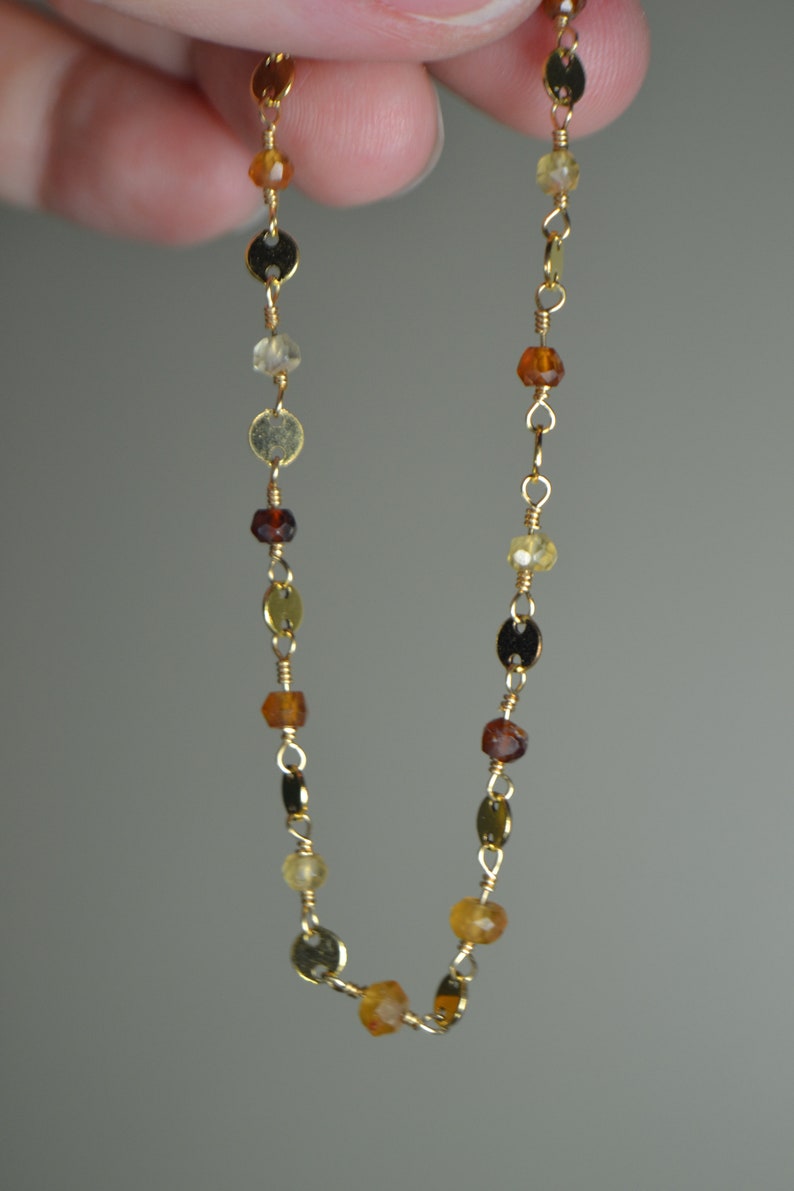 May include: Delicate gold necklace featuring a series of small, colorful gemstones and gold-colored disc accents. The gemstones vary in shades of amber, brown, and yellow, strung together with fine gold wire.
