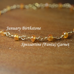 May include: A gold-coloured chain bracelet featuring orange Spessartine garnet beads. The image includes the text "January Birthstone" and "Spessartine (Fanta) Garnet". The bracelet is presented on a wooden surface.
