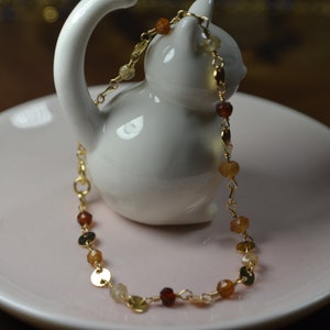 May include: A delicate gold necklace with a variety of gemstones in shades of orange, yellow, and brown. The necklace is displayed on a white ceramic cat figurine, resting on a pink plate.