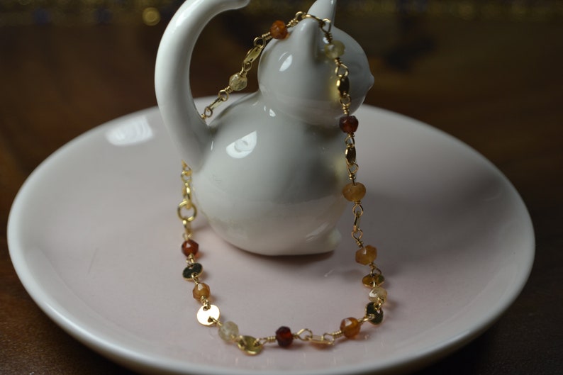 May include: A delicate gold necklace with a variety of gemstones in shades of orange, yellow, and brown. The necklace is displayed on a white ceramic cat figurine, resting on a pink plate.