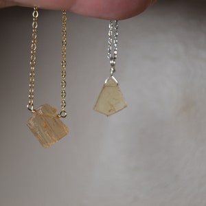 May include: Two necklaces with light orange-brown crystal pendants. One necklace has a gold chain, and the other has a silver chain. The pendants have a rough, natural appearance. The background is a soft, neutral color.