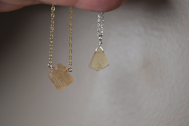 May include: Two necklaces with light orange-brown crystal pendants. One necklace has a gold chain, and the other has a silver chain. The pendants have a rough, natural appearance. The background is a soft, neutral color.