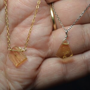 May include: Two necklaces with amber-colored pendants. One necklace has a gold chain, and the other has a silver chain. The pendants are irregularly shaped, with a faceted appearance. The necklaces are displayed against a skin-toned background.