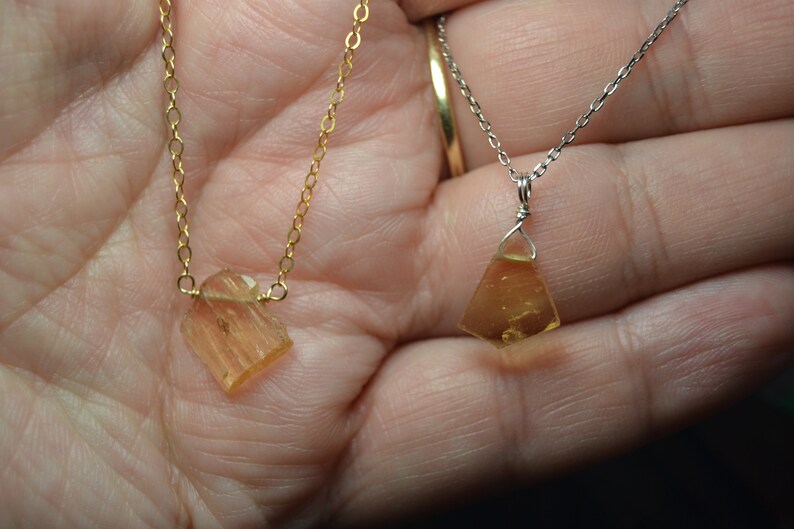 May include: Two necklaces with amber-colored pendants. One necklace has a gold chain, and the other has a silver chain. The pendants are irregularly shaped, with a faceted appearance. The necklaces are displayed against a skin-toned background.