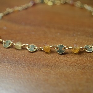 May include: A delicate gold-toned bracelet featuring small, round gold accents and colorful gemstones. The gemstones are in shades of yellow, orange, and brown. The bracelet is displayed on a wooden surface.