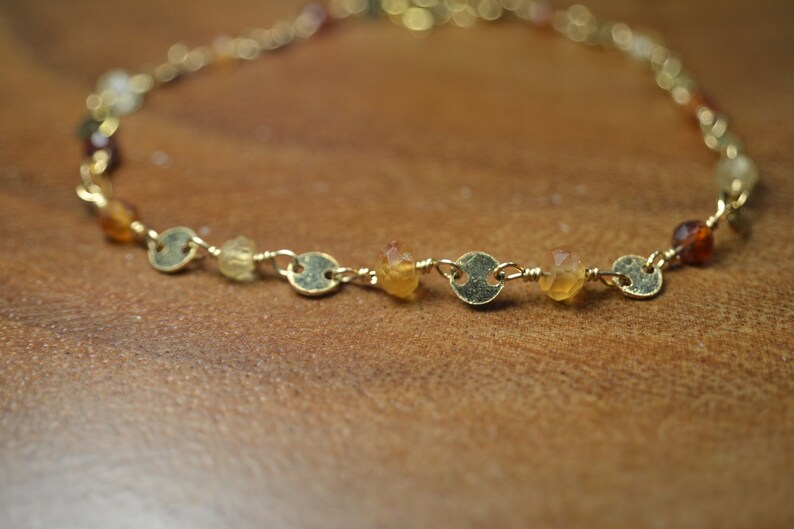 May include: A delicate gold-toned bracelet featuring small, round gold accents and colorful gemstones. The gemstones are in shades of yellow, orange, and brown. The bracelet is displayed on a wooden surface.