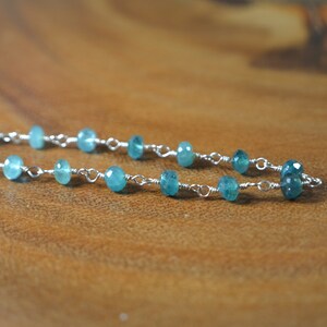 May include: A delicate necklace featuring small, faceted, teal-colored gemstones strung on a silver chain. The gemstones are evenly spaced, creating a subtle sparkle. The necklace is displayed on a wooden surface.