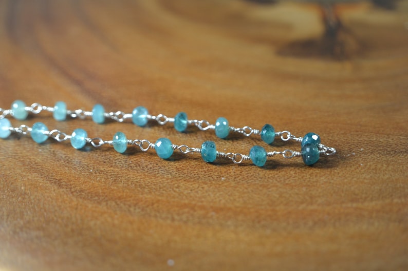 May include: A delicate necklace featuring small, faceted, teal-colored gemstones strung on a silver chain. The gemstones are evenly spaced, creating a subtle sparkle. The necklace is displayed on a wooden surface.