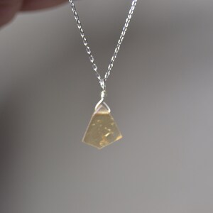 May include: A silver chain necklace with a small, irregularly shaped, amber-colored pendant. The pendant is suspended from a delicate silver chain, creating a minimalist and elegant design. The necklace is shown against a soft, neutral background.