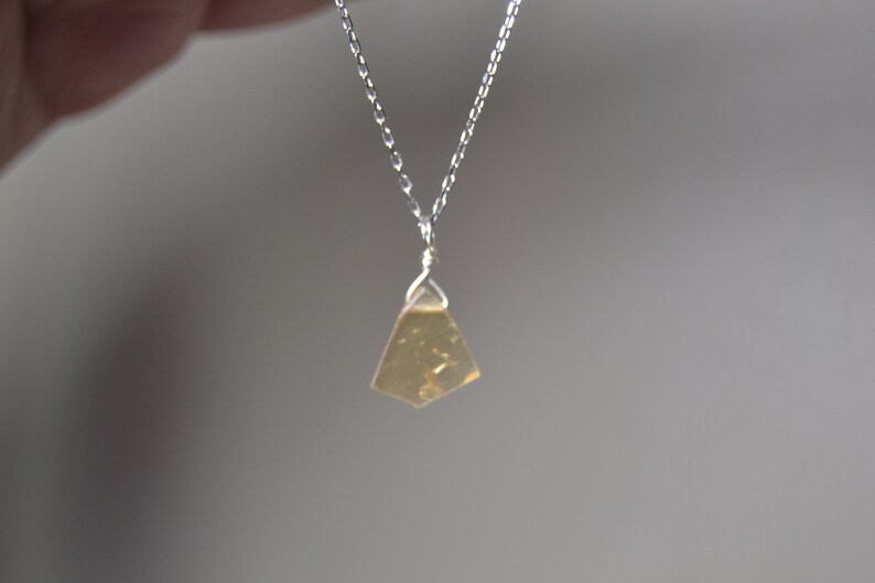 May include: A silver chain necklace with a small, irregularly shaped, amber-colored pendant. The pendant is suspended from a delicate silver chain, creating a minimalist and elegant design. The necklace is shown against a soft, neutral background.