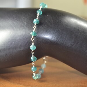 May include: A delicate bracelet featuring small, round, teal-colored beads strung on a silver-colored chain. The beads are evenly spaced along the chain, creating a simple yet elegant design. The bracelet is shown on a dark surface.