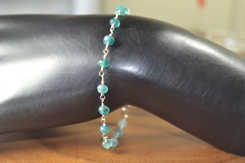 May include: A delicate bracelet featuring small, round, teal-colored beads strung on a silver-colored chain. The beads are evenly spaced along the chain, creating a simple yet elegant design. The bracelet is shown on a dark surface.