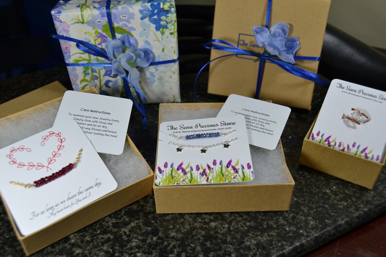 May include: An assortment of jewelry items, including necklaces and rings, presented in gift boxes. Some boxes are wrapped in floral paper and tied with blue ribbon. Each box contains a card with care instructions and the shop's name.