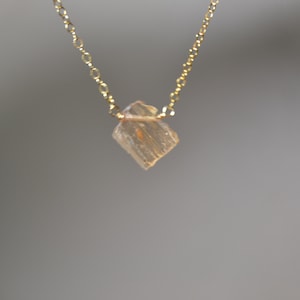 May include: A delicate gold necklace featuring a small, faceted, light-colored crystal pendant. The pendant is a square shape with a slightly rough texture. The chain is a fine, linked design. The necklace is displayed against a neutral background.