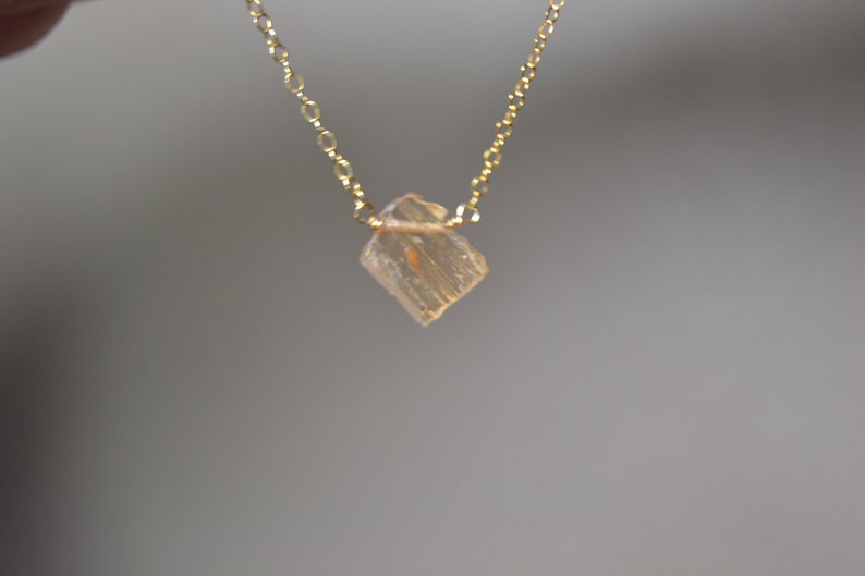 May include: A delicate gold necklace featuring a small, faceted, light-colored crystal pendant. The pendant is a square shape with a slightly rough texture. The chain is a fine, linked design. The necklace is displayed against a neutral background.