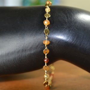 May include: Gold-toned bracelet featuring a delicate chain interspersed with small, round, and flat gold discs. The bracelet also includes a series of small, orange-toned gemstone beads. The bracelet is displayed against a dark background.