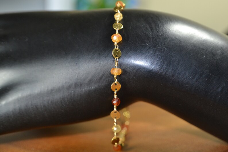May include: Gold-toned bracelet featuring a delicate chain interspersed with small, round, and flat gold discs. The bracelet also includes a series of small, orange-toned gemstone beads. The bracelet is displayed against a dark background.