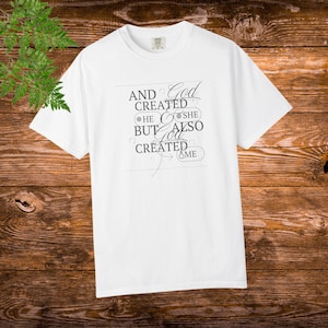 May include: White t-shirt featuring the text "AND GOD CREATED HE SHE BUT ALSO GOD CREATED ME" in black, stylish lettering. The shirt has a crew neck and short sleeves. The background is a wooden surface.