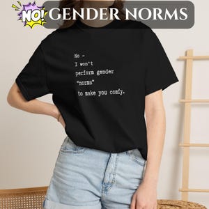 Puede incluir: Camiseta negra con el texto "No - I won't perform gender 'norms' to make you comfy." Las palabras "NO! GENDER NORMS" están en la parte superior en un bocadillo. La camiseta es una prenda informal.